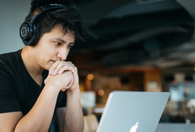 A young man with a black shirt sat at a desk looking at his MacBook with black headphones on