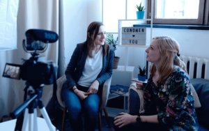 Two women in a room, in front of a camera having a conversation.